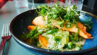 A colorful dish featuring a cooked salmon fillet topped with fresh greens, sitting on a bed of mixed vegetables like carrots and peas in a creamy sauce. The meal is presented in a dark blue bowl on a green table, with glasses and cutlery nearby.